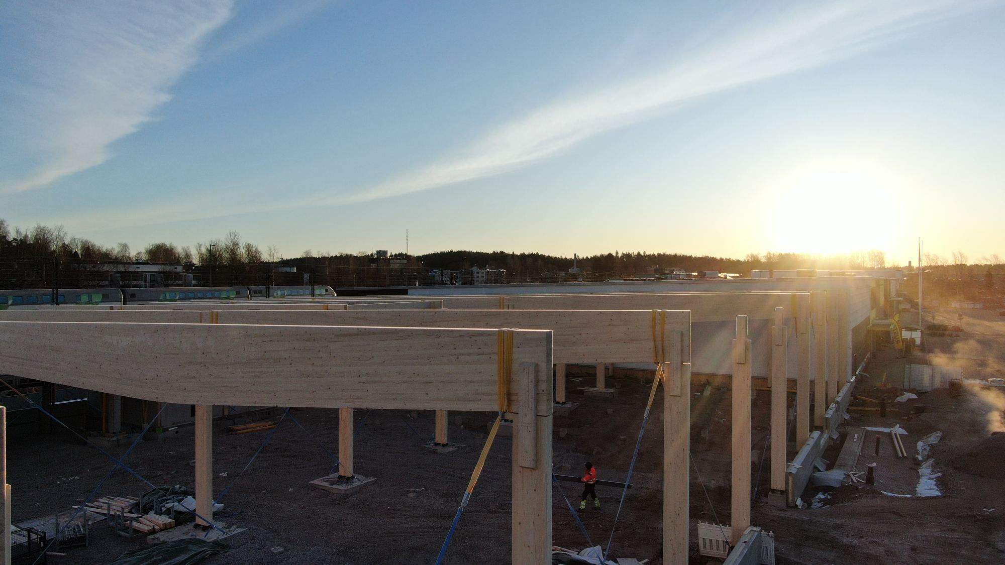 Liimapuupalkit ja -pilarit ilta-auringossa Kirkkonummen Tähden työmaalla, rakentaja palkkien alla